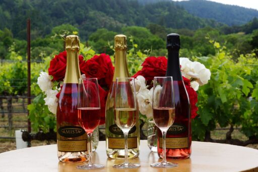 three bottles of Amista Vineyards sparkling wine with glasses of sparkling wine and flowers and vineyards in the background