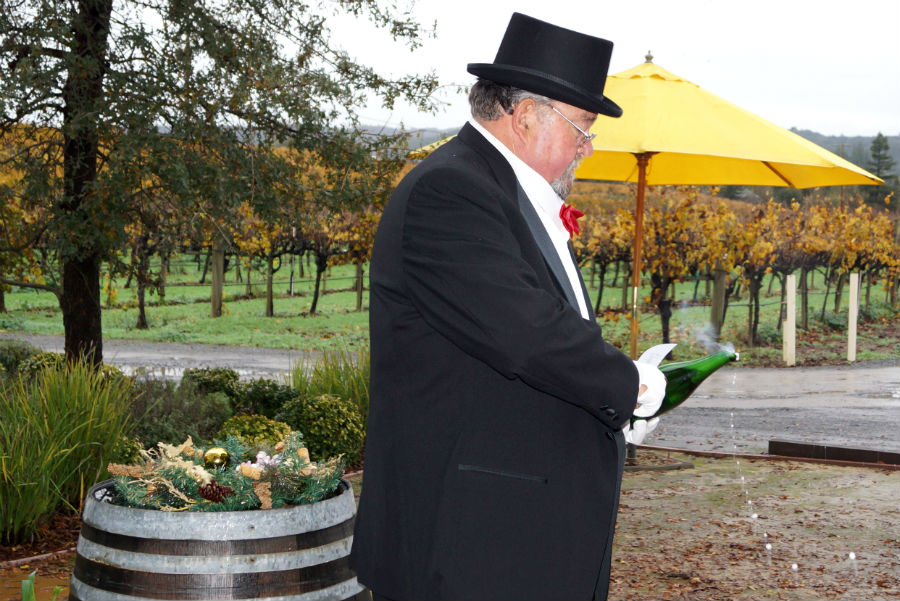Amista Vineyards Co-founder Mike Farrow in tux, top hat and red bowtie, with bottle of sparkling wine standing in the vinyards with a yellow umbrella in the background