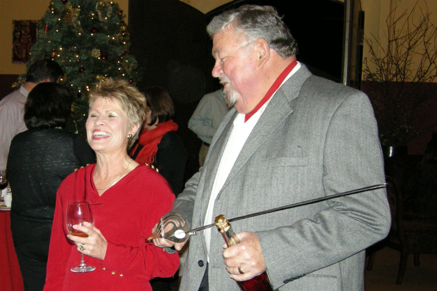 Amista Vineyards Co-founders Vicky Farrow, smiling with glass of wine and Mike Farrow smiling with bottle of wine and sword in his hand