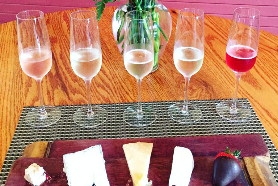 Five glasses of Amista Sparkling wines from pale the pink to cranberry color a board of cheese wedges and a chocolate covered strawberry