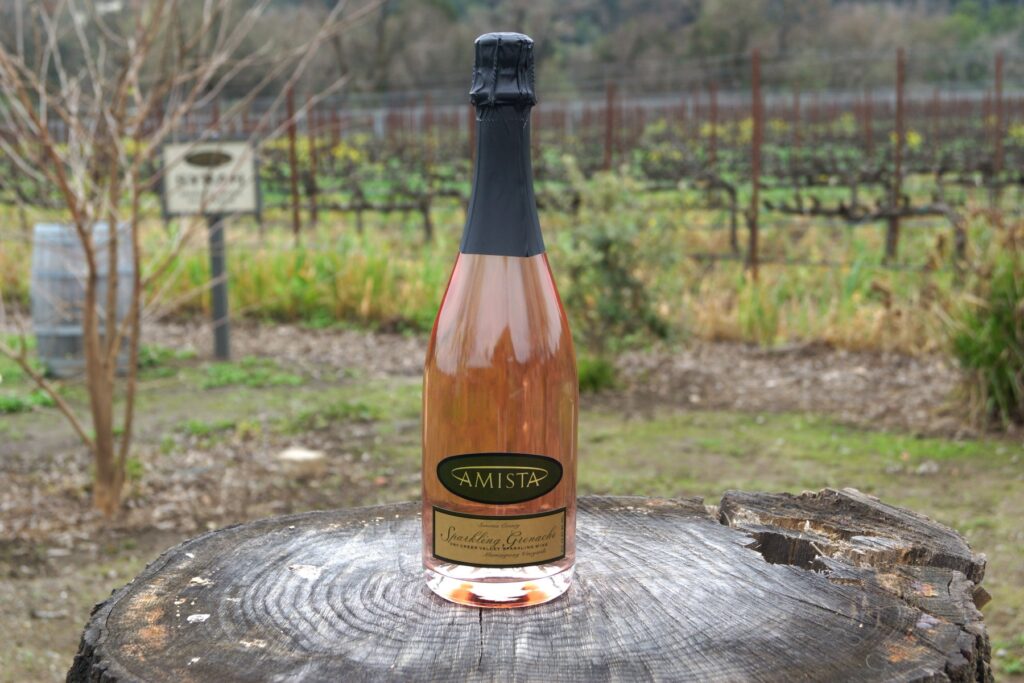 Amista Vineyards Sparkling Grenache sitting on a tree stump in the vineyards with a barrel and sign with the word Syrah in the background
