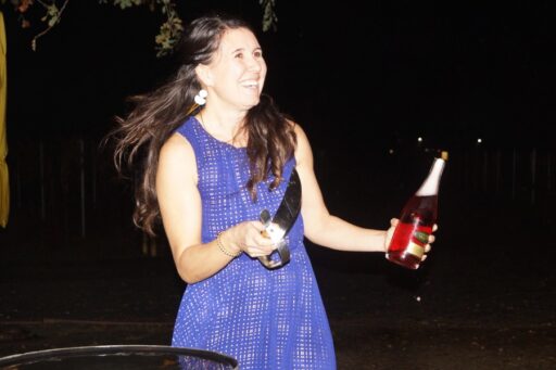 Ashley Herzberg, Amista Winemaker, smiling in purple dress holding a bottle of cranberry colored sparkling wine and a saber