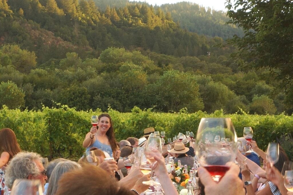 People seated at long table among the vines raising their glasses at Amista Dinner in the Vineyard
